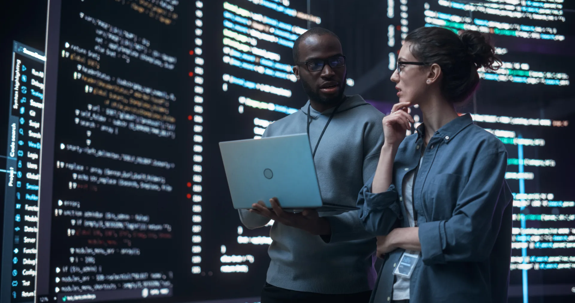 Two colleagues discuss code on a laptop in front of tall screens filled with highlighted source code.