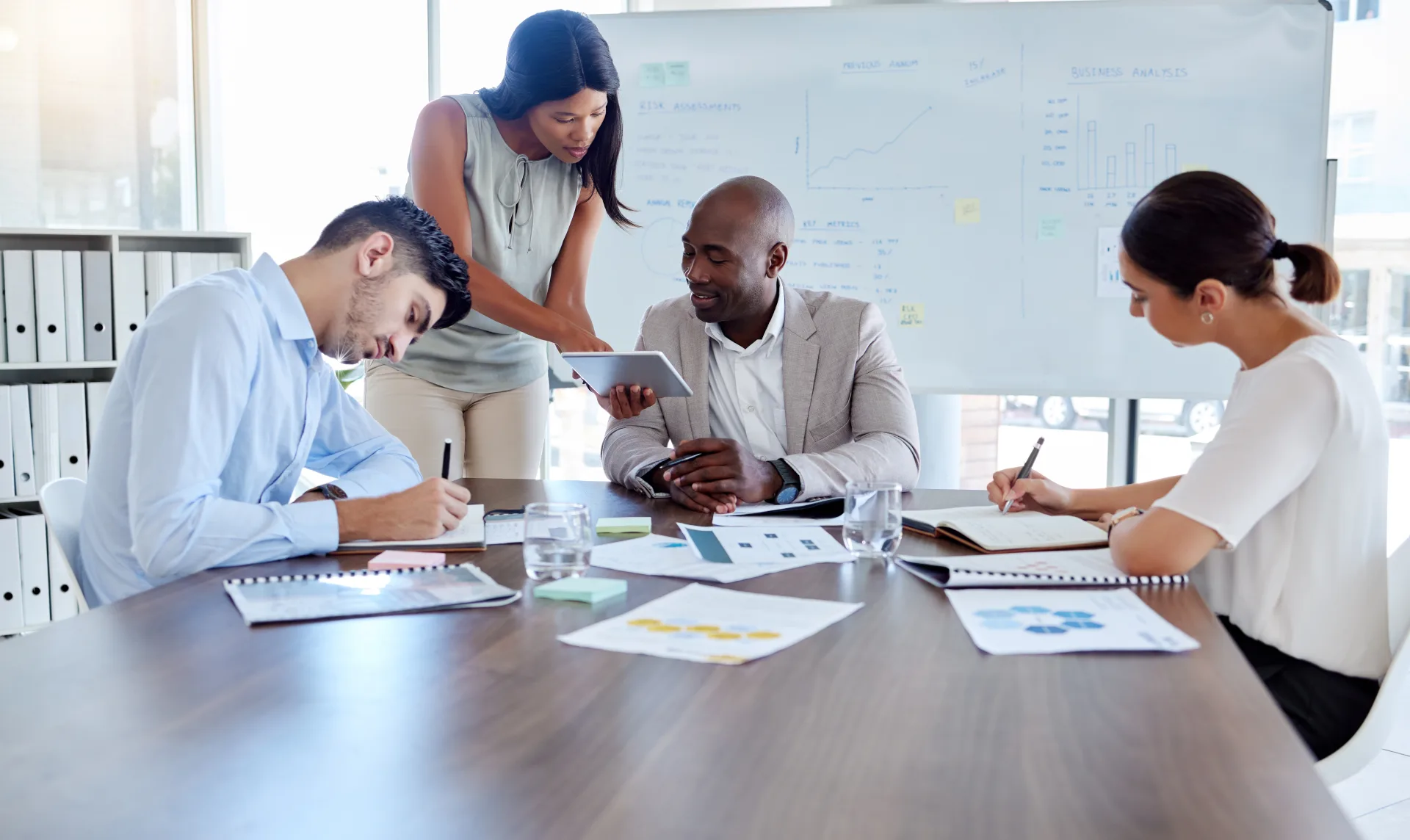 Four colleagues review printed charts and a tablet at a table, beside a whiteboard headed Business Analysis.