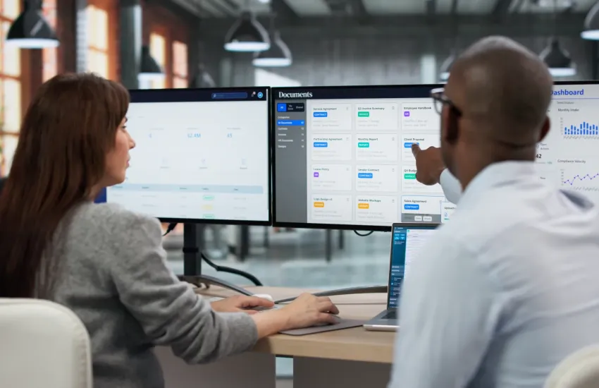 Two colleagues review a document management dashboard of contracts and invoices on dual monitors in an open-plan office.