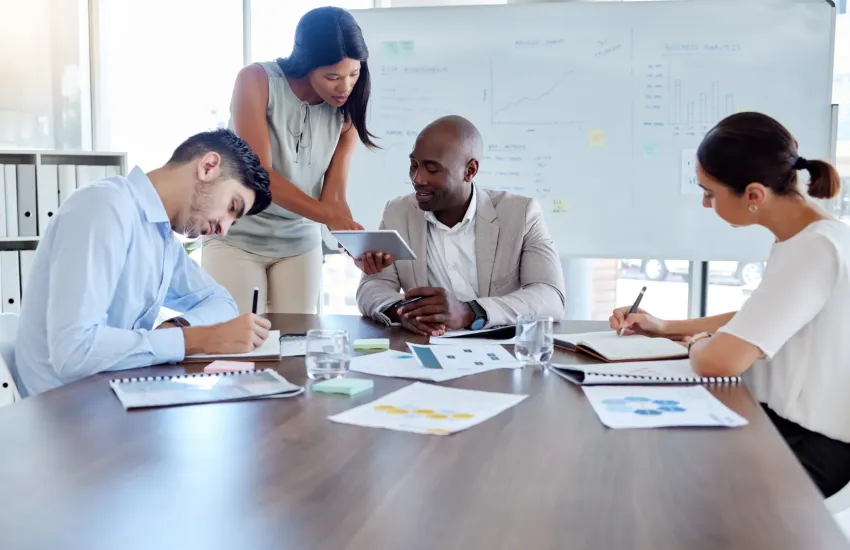 Four colleagues review printed charts and a tablet at a table, beside a whiteboard headed Business Analysis.