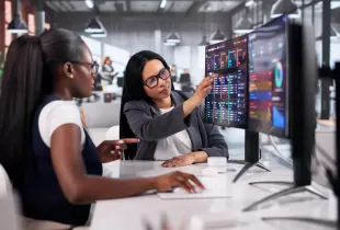 Two women professionals collaborate at a desk; reviewing colorful data dashboards on a large monitor in a modern office.