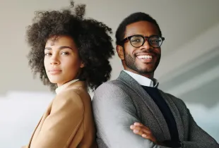 Two professionals in business casual stand back to back, one smiling at the camera in a bright office.