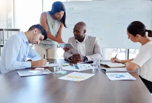 Four colleagues review printed charts and a tablet at a table, beside a whiteboard headed Business Analysis.