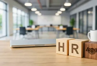 Wooden blocks spelling HR with a handshake icon on an administrative office desk; with a laptop; coffee mug; and open workspace in the background.